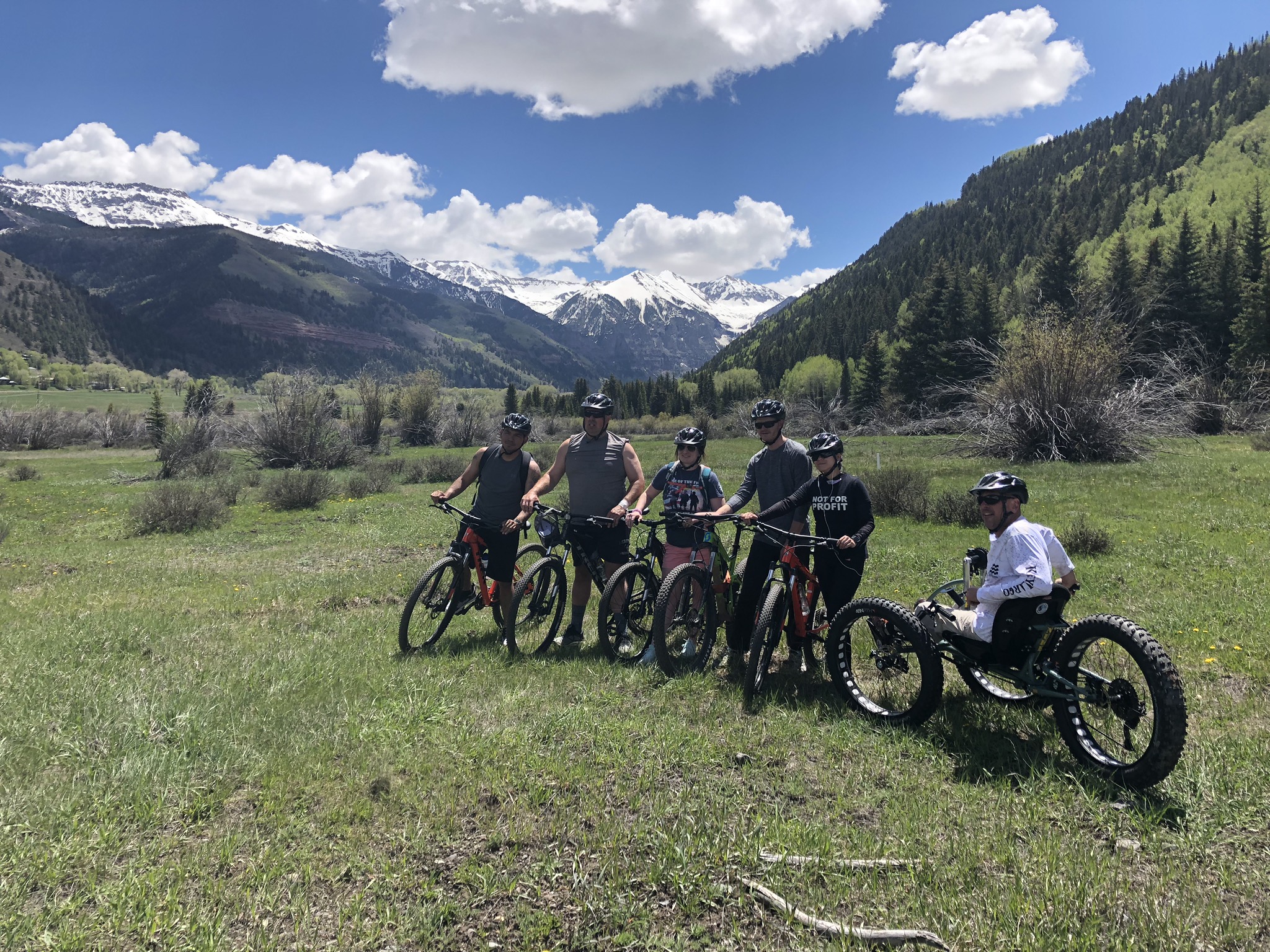 A group of six people wearing helmets, posing with their bicycles in a grassy field with mountains in the background. One person is on a recumbent bike. The sky is clear and blue with scattered clouds, and the surrounding trees are lush and green—perfect weather for cycling.