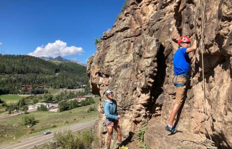 Two people are engaging in rock climbing on a sunny day. One, in a blue shirt and red helmet, is scaling the rock face while the other, dressed in a gray shirt and helmet, is belaying. A road winds through the green landscape with mountains visible in the background, making for perfect adventure activities.