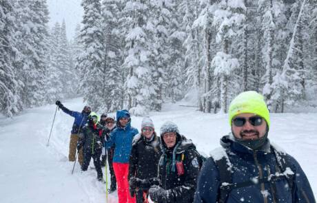 A group of people standing on a snowy path in a forest, dressed in winter gear including hats, jackets, and gloves. Snow is falling lightly and the trees around them are covered in snow. One person in the foreground wears a neon green hat and sunglasses, ready for their snowshoeing adventure.