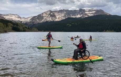 A group of people paddleboarding on a lake with snow-capped mountains and a forest in the background. Some participants are using adapted paddleboards, including one person in a wheelchair on a paddleboard. The sky is partly cloudy, making for an inspiring scene of inclusive sports.