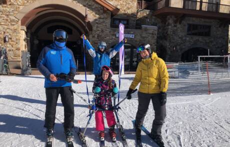 Four people dressed in winter gear are enjoying a ski day. One person, in a yellow jacket, is on the right, while another in blue is on the left. Two individuals are assisting a seated skier in the center. In the background, snow-covered ground and buildings set the serene alpine skiing scene.