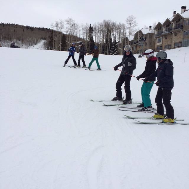 A group of six skiers, dressed in winter gear and helmets, stand on a snowy slope with ski poles. They are positioned in a line across the slope, preparing to ski as part of TASP training. In the background, there are bare trees and chalet-style buildings.