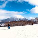 A snowy landscape with several people skiing on a slope under a bright blue sky with scattered clouds. In the background, a snow-capped mountain rises above a dense forest, partially shrouded in mist.