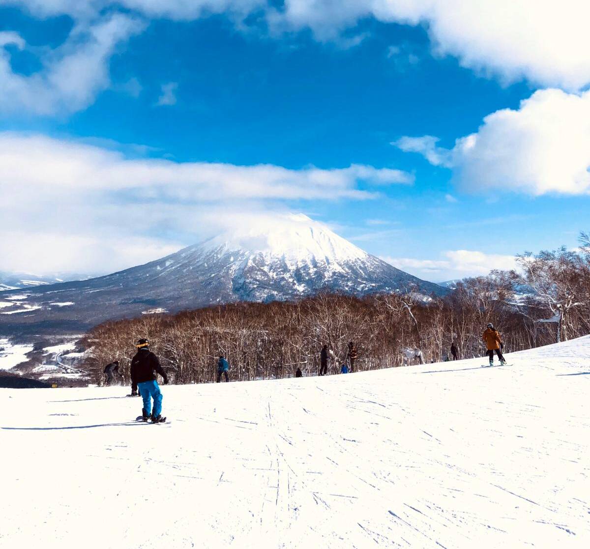 A snowy landscape with several people skiing on a slope under a bright blue sky with scattered clouds. In the background, a snow-capped mountain rises above a dense forest, partially shrouded in mist.