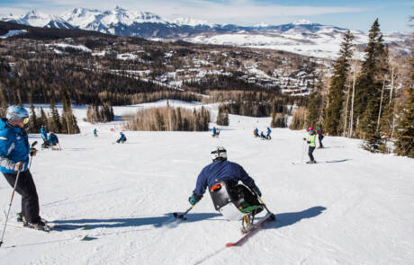 People are skiing down a snowy mountain slope with a scenic view of snow-capped mountains and a forested landscape in the background. A skier in adaptive equipment is in the foreground, showcasing the inclusivity of winter programs, while other skiers are seen scattered down the slope.