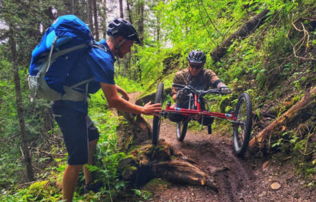 A man assists another navigating a forest trail on an adaptive handcycle, showcasing the spirit of adaptive sports. Surrounded by dense foliage, they maneuver around a large fallen tree, focusing intently as teamwork helps them tackle the challenging terrain.