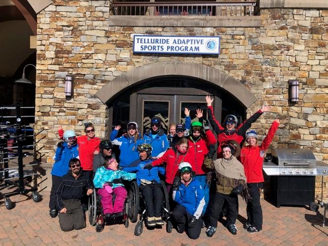 A group of people poses in front of a building with a sign above reading "Telluride Adaptive Sports Program." They appear to be skiing enthusiasts, some in wheelchairs and others in skiing gear. Everyone is smiling and raising their arms joyfully.