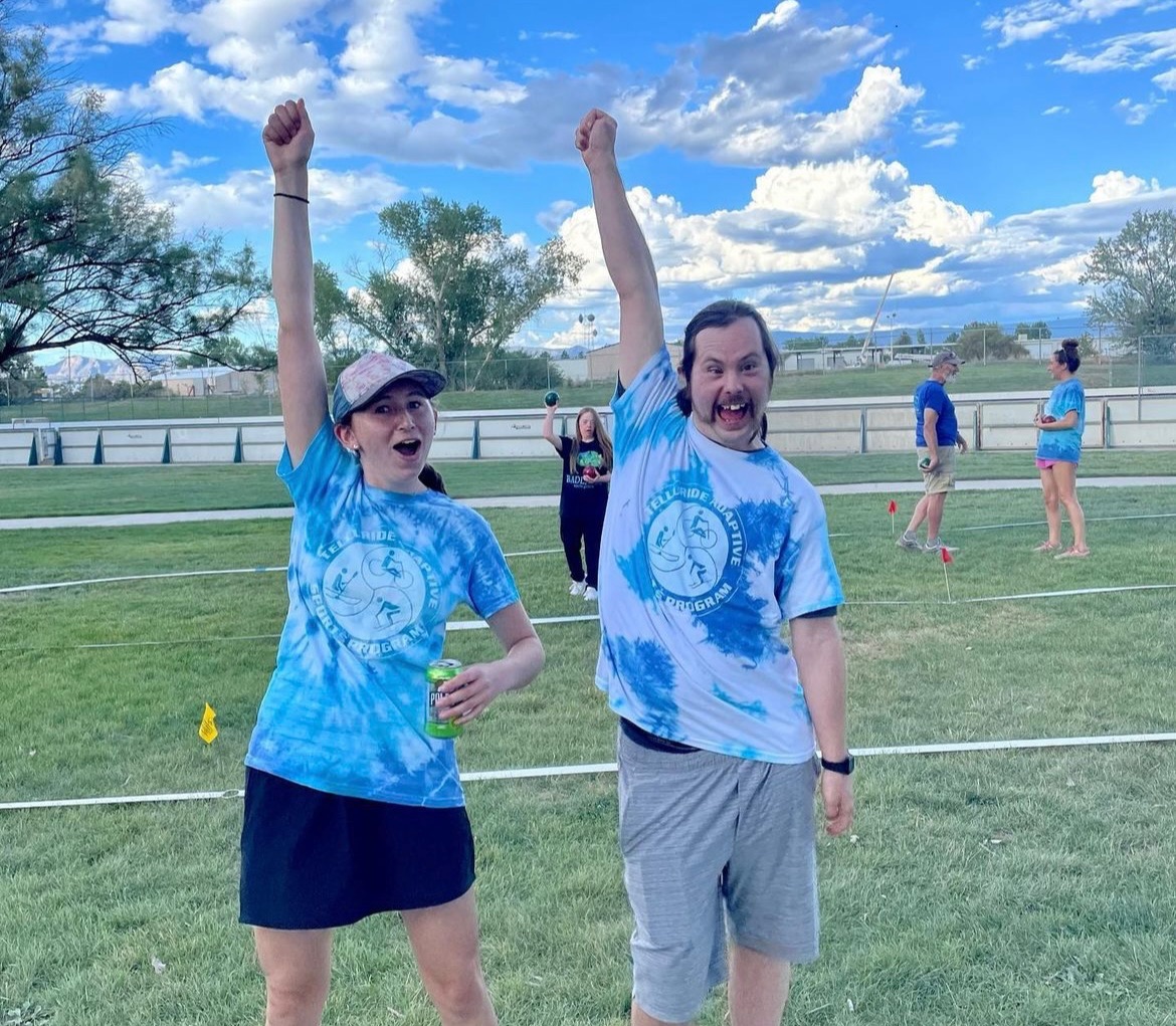 Two ecstatic individuals stand on a grassy field with their arms raised in victory, embodying the joy of summer fun. Both wear tie-dye blue and white T-shirts, one holding a drink and the other cheering animatedly. A few other people are seen in the background under a bright blue sky with fluffy clouds.