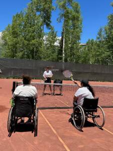 Two individuals in wheelchairs are having fun playing tennis on a clay court during a sunny summer day. One person is on the left, the other on the right, both facing the net. A coach stands behind the net, holding a tennis racket. Lush green trees and a clear blue sky form the background.