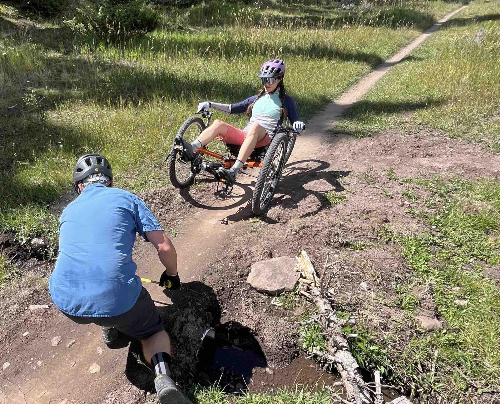 A person in a blue shirt is placing a rock into a hole on a dirt path, while another individual in a recumbent bicycle, both members of the Mountain Club, waits behind them. Both are wearing helmets. The scene is set in a grassy outdoor area with a clear sky.