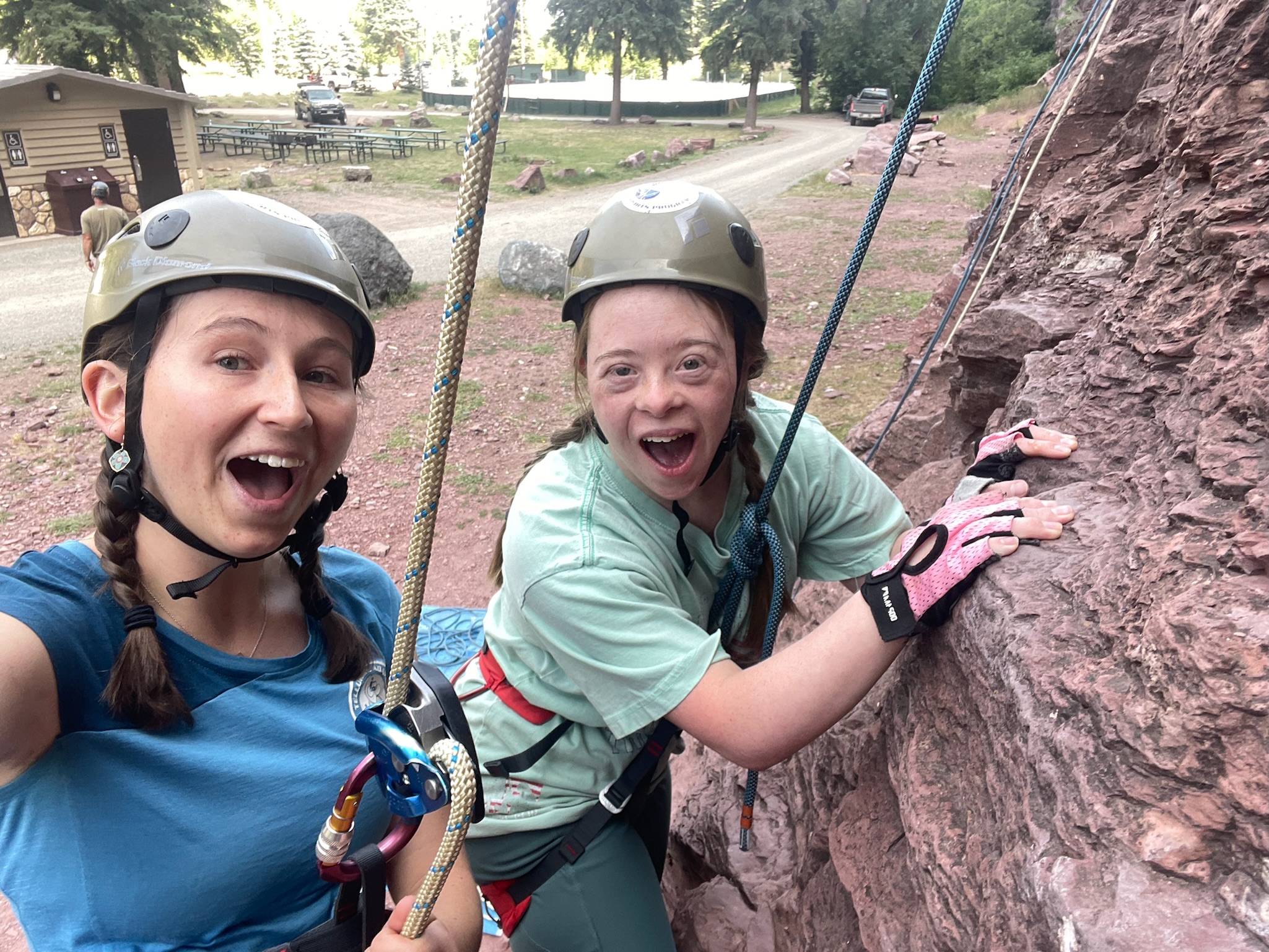 Two people wearing helmets are rock climbing outdoors as part of exciting program activities. One smiles at the camera while the other focuses on climbing the rocky surface. They are secured with harnesses and ropes, enjoying their adventure. A building and picnic tables can be seen in the background.