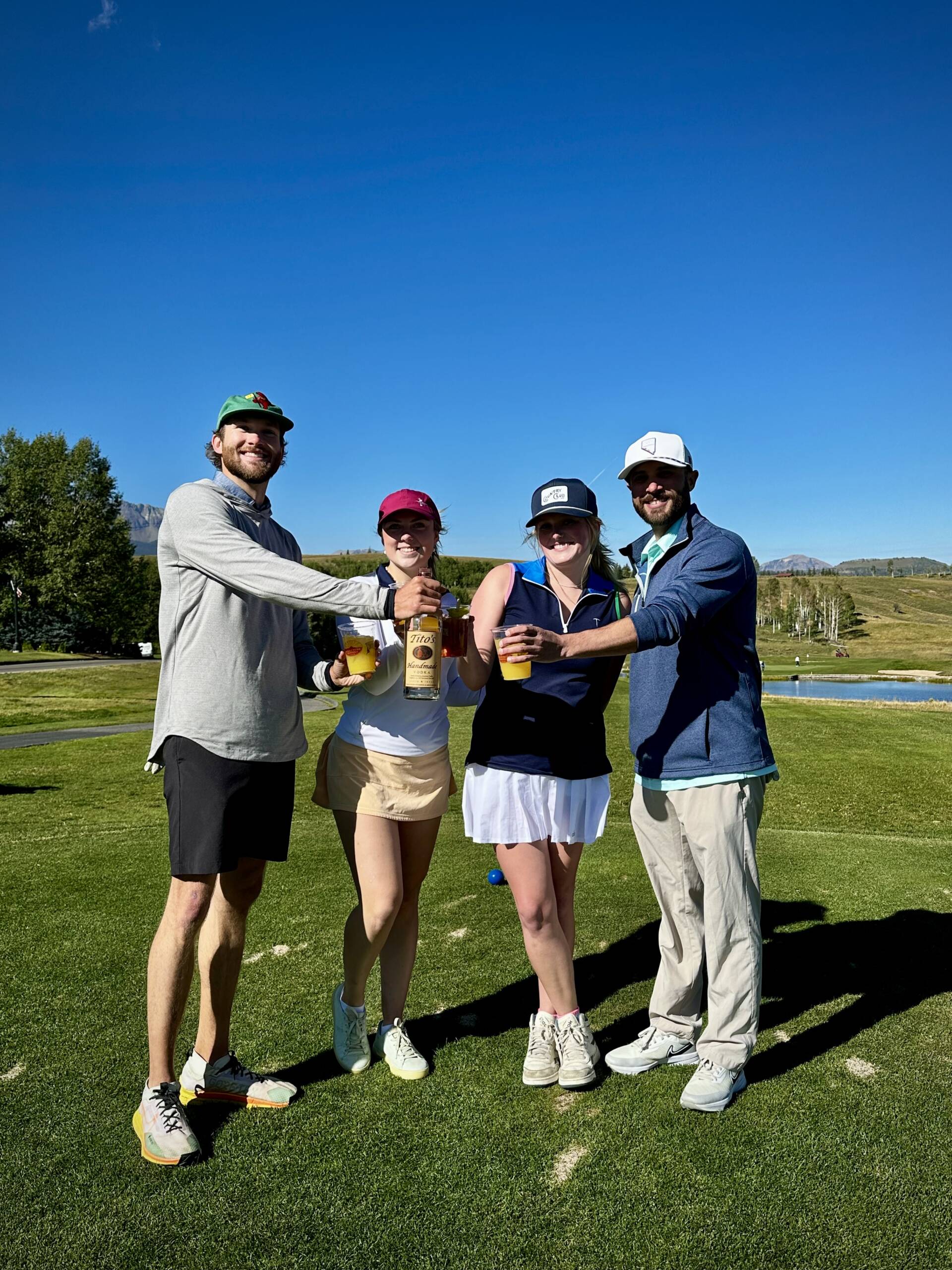 Four people standing on a golf course, smiling and holding drinks at a fundraising event. Dressed in casual golf attire, they seem to be enjoying the sunny day outdoors. Trees and a clear blue sky provide the perfect backdrop for this engaging gathering.