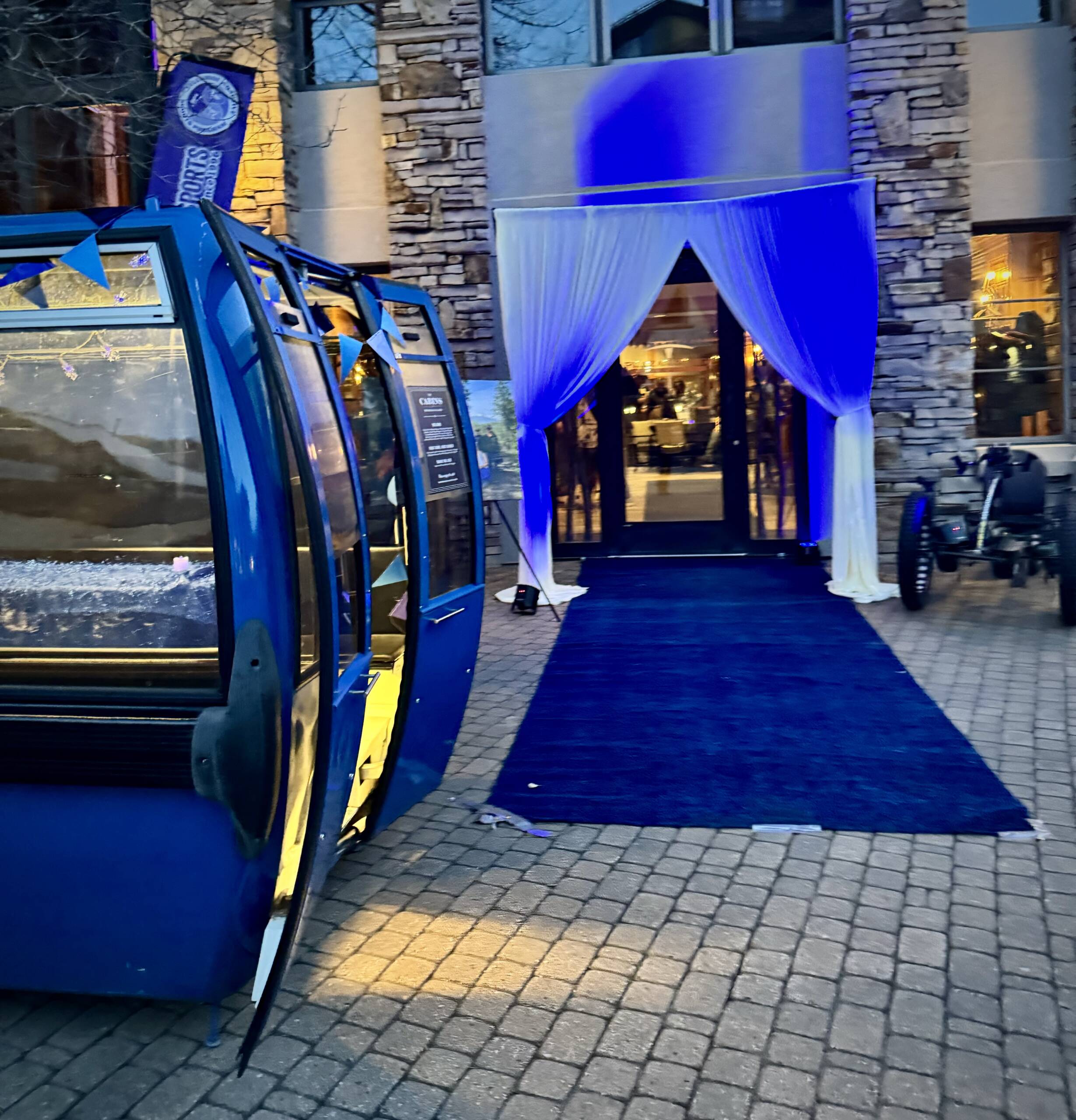 A blue gondola cabin is placed on a cobblestone path next to a fundraising event entrance. The entrance features white drapes and blue lighting, leading into a building with stone walls and large windows. A bicycle is parked nearby.