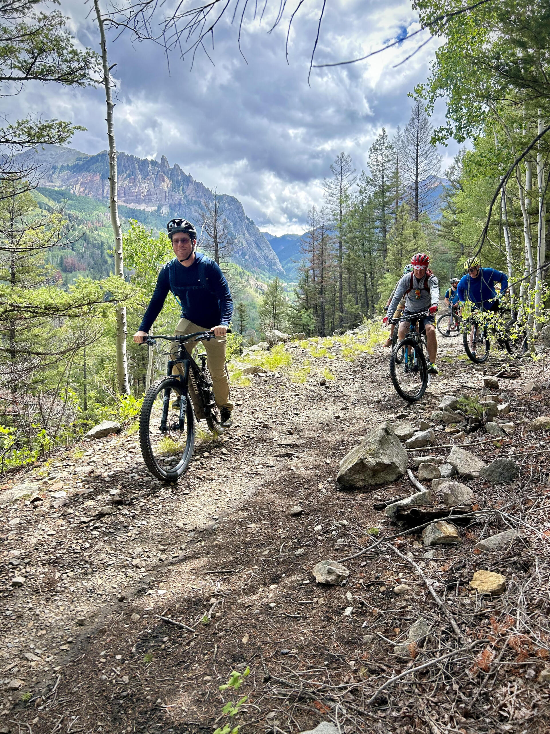 Three people mountain biking on a rugged forest trail, part of exciting program events. Rocky terrain and tall trees stretch towards majestic mountains under a partly cloudy sky, evoking a thrilling sense of adventure and outdoor exploration.