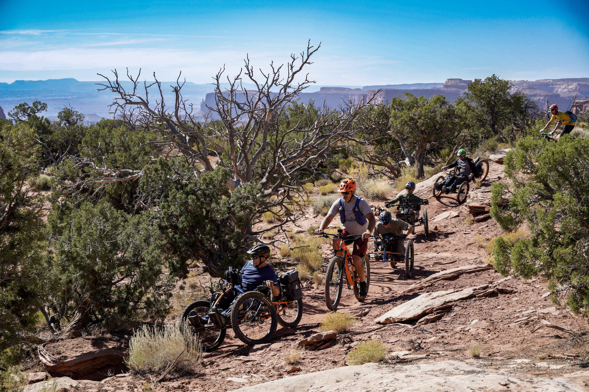 A group of cyclists, including those on adaptive bikes, participates in an adventurous program as they traverse a rocky desert trail. Events unfold under the clear blue sky, with sparse vegetation and distant mesas visible in the backdrop.