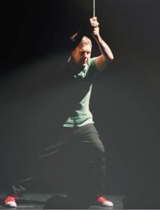 A person in a green shirt and red shoes stands on a dimly lit stage, gripping a rope above their head. The light highlights their focused expression and dynamic pose. Amidst the shadows, the scene feels like a dramatic movie screening with all eyes drawn to them in the spotlight.