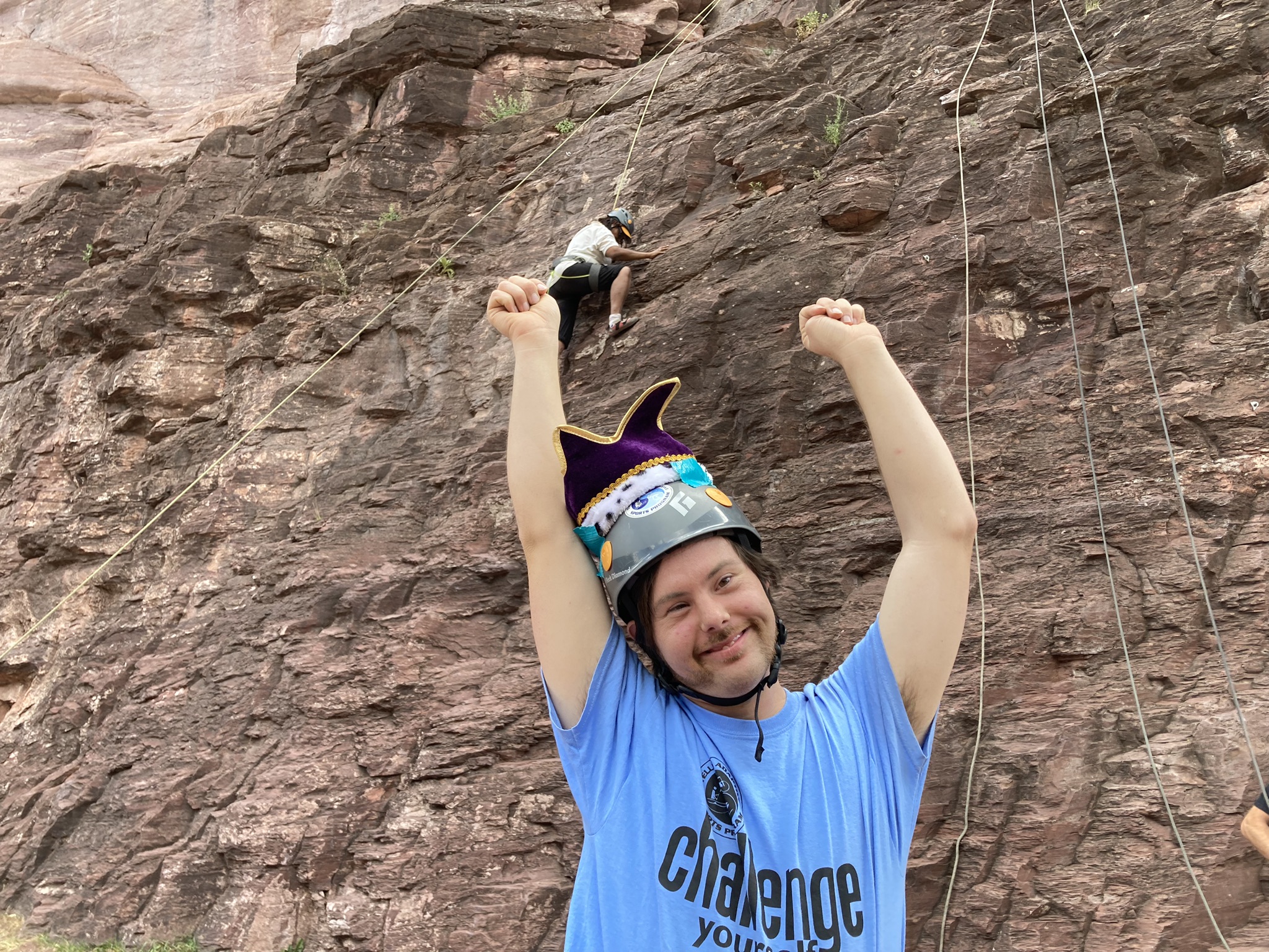 A person with a costume knight hat on his climbing helmet and blue shirt raises their arms and smiles at the camera, while another person climbs a rocky cliff in the background.
