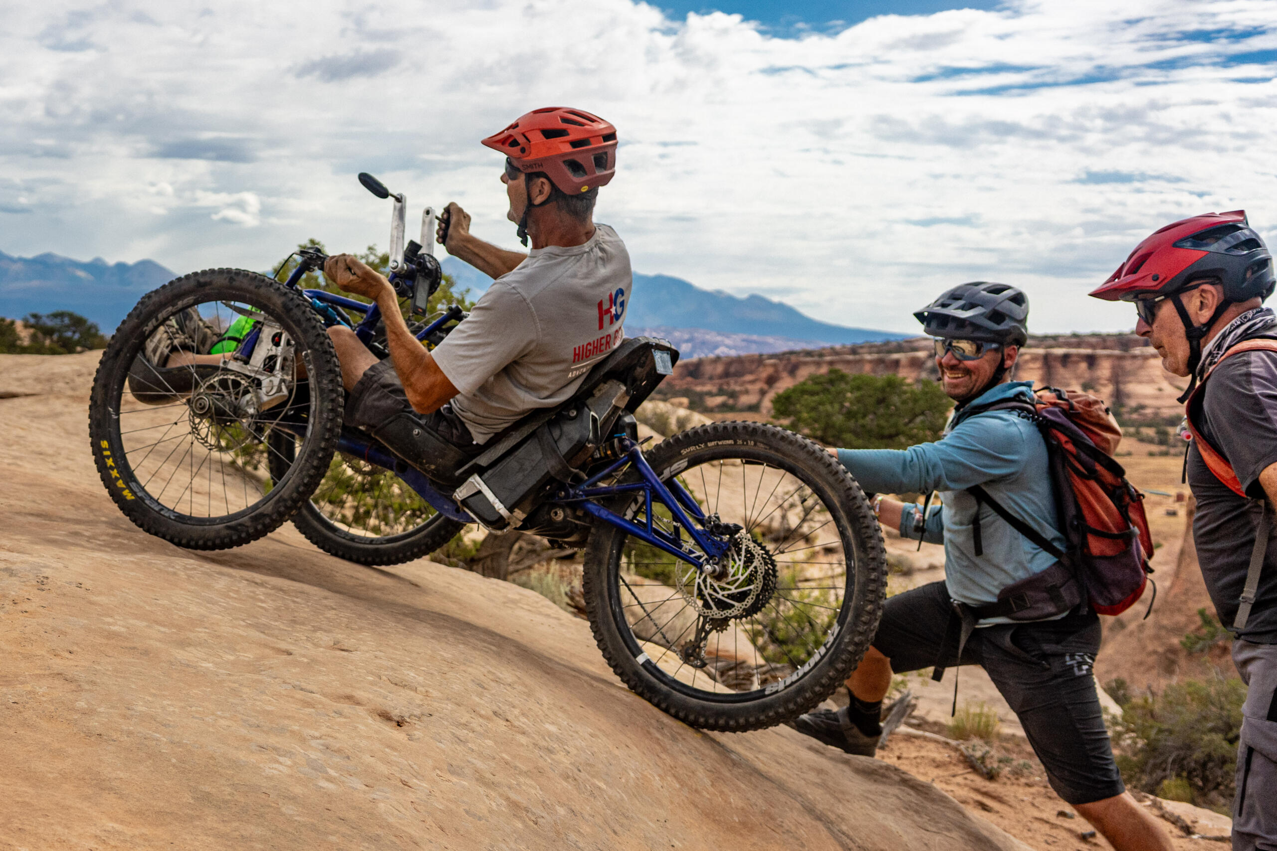 A man in a handcycle is helped up a rocky slope by two people in helmets and outdoor gear, with a scenic desert landscape in the background.